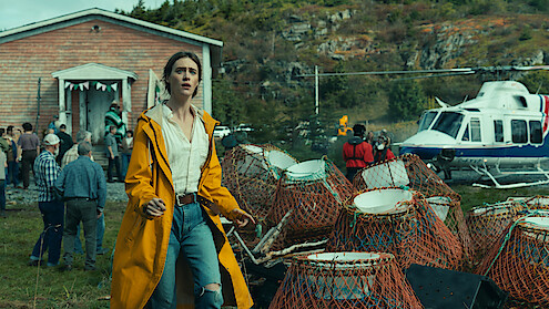 Woman in yellow raincoat stands near lobster traps, outdoors by a crowd and a helicopter, with a wooden building and forested rocky hill in the background, suggesting a coastal rural setting.