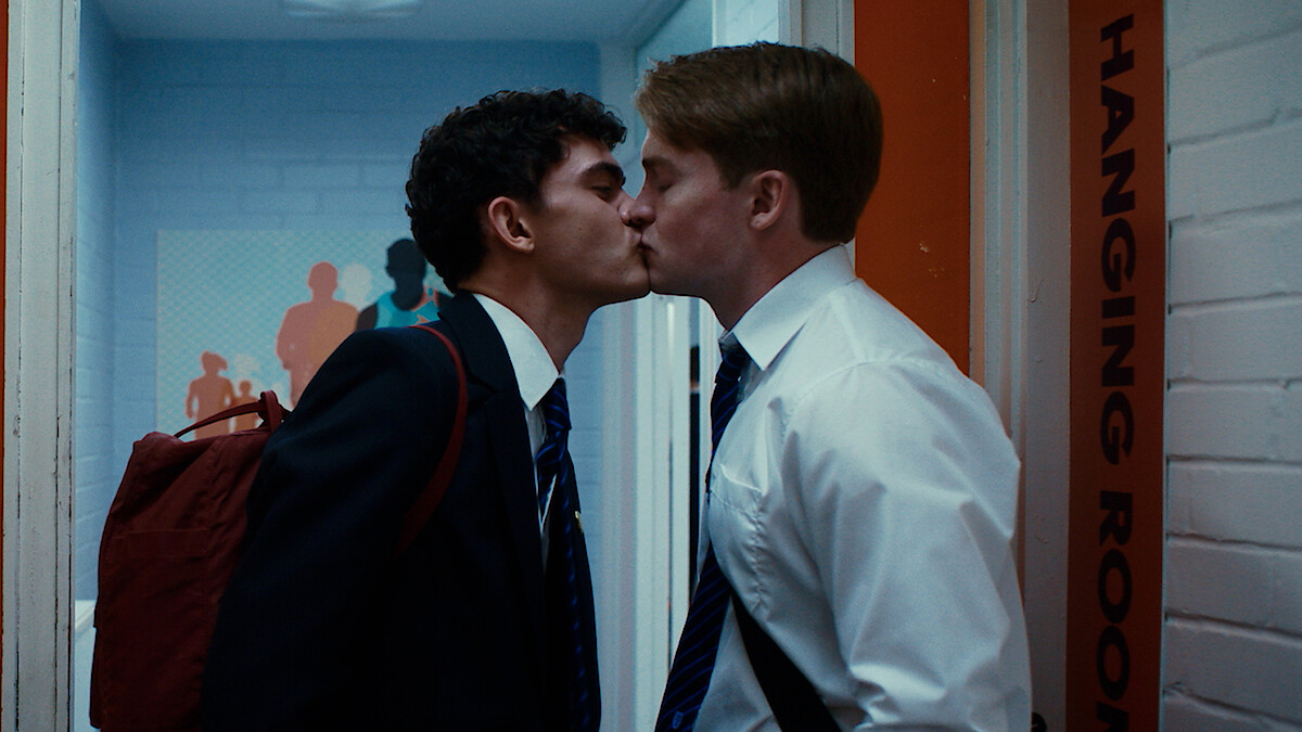 Two young men in school uniforms kiss in a hallway next to a door labeled "HANGING ROOM." One wears a backpack, and the background features colorful wall art and a white brick wall.
