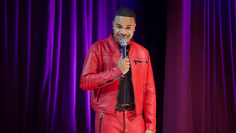 A comedian in a bright red leather outfit performs stand-up on stage, holding a microphone, with purple stage curtains in the background and stage lighting.