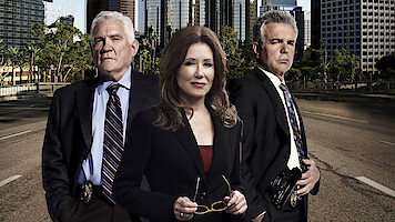 Three serious-looking detectives in suits stand on an empty city street with tall buildings and trees in the background, suggesting an urban law enforcement setting.