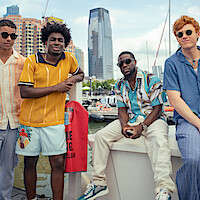 Four men in stylish summer outfits pose at a marina with boats, water, and tall city buildings in the background, suggesting a relaxed, urban waterfront setting.
