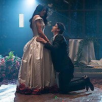 A man in a suit kneels before a woman in a white gown with a blood-stained hem. They are in a dimly lit, decorated indoor setting with flowers and greenery, creating a dramatic and tense atmosphere.