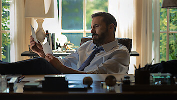 A man in a shirt and tie sits at a desk in a sunlit office, reading a document with his feet up. The office has large windows, books, and various desk items visible in the background.