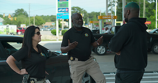Two uniformed police officers stand in a gas station parking lot talking to a man in a black hooded sweatshirt, black pants and a black mask.
