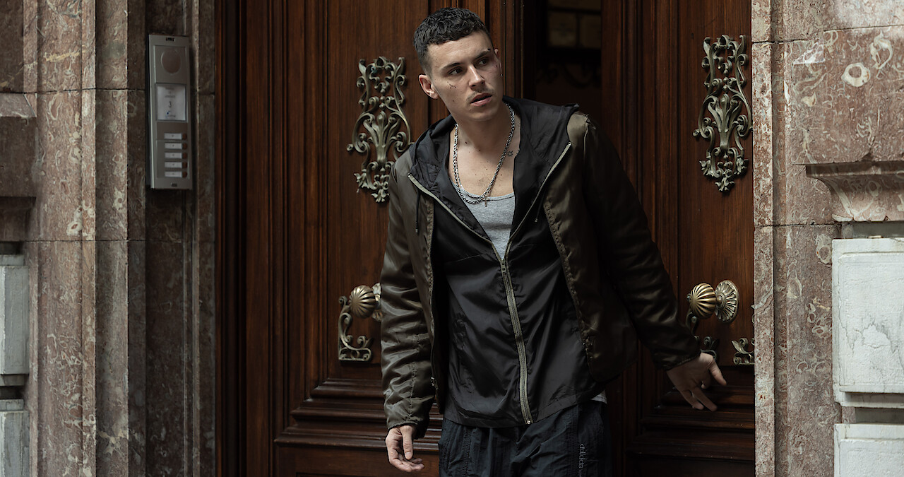 A man wearing a black sweatshirt and brown jacket stands in an open doorway.