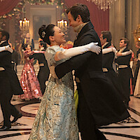A couple in formal period costumes dances in a grand, elegant ballroom with chandeliers, red drapes, and other couples waltzing in the background during a lavish ball.