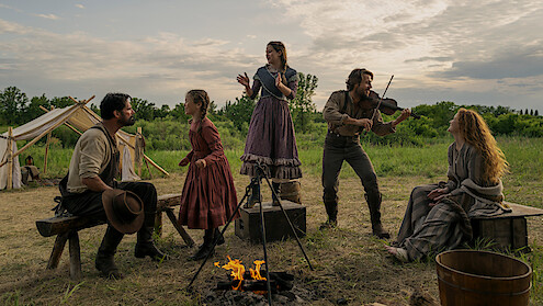 Five people in pioneer-era clothing gather around a campfire in an open field with tents. One plays a violin, others sing or listen, creating a lively, outdoor, historical family campsite scene at sunset.