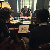 Three people in an office: a man behind a desk and two others sitting across, showing sketches. The setting has classic decor, sunlight filtering through blinds, and papers and drawings spread out, suggesting a serious discussion or review.