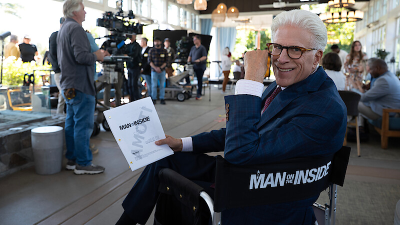Smiling man in a suit holding a script, sitting on a director’s chair labeled "Man on Inside" on a bustling film set with crew, cameras, and lighting equipment in a modern, well-lit indoor space.