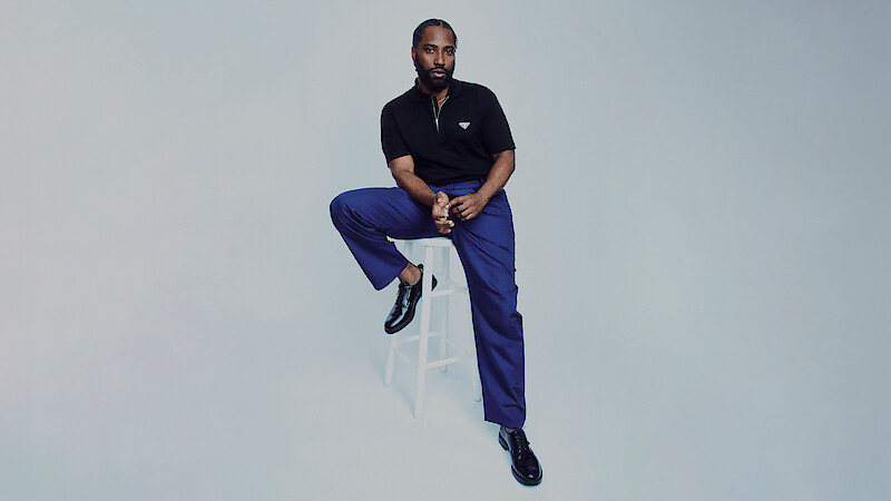 John David Washington sits on a chair and stretches his leg out against a grey background.