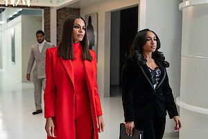 Crystle Stewart as Mallory and Taylor Polidore Williams as Kimmie in business attire, one in a red suit and the other in a black suit, walk confidently down a modern office hallway, with a man in a grey suit walking behind them.