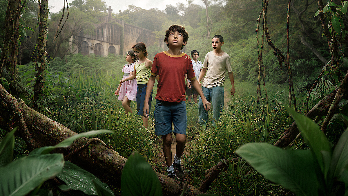 A group of children walk through dense green jungle foliage, looking around curiously. In the background, a large, old, abandoned stone building stands partially hidden by mist and trees, creating an adventurous and mysterious atmosphere.