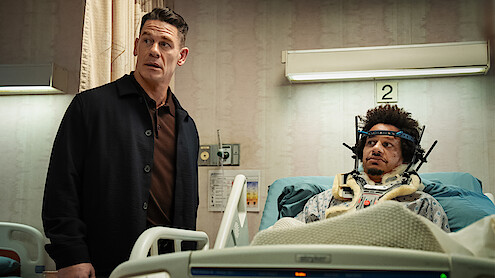 A man stands next to a hospital bed in a room, while another man lies in the bed wearing a neck brace and head immobilizer, suggesting a medical emergency or recovery setting.