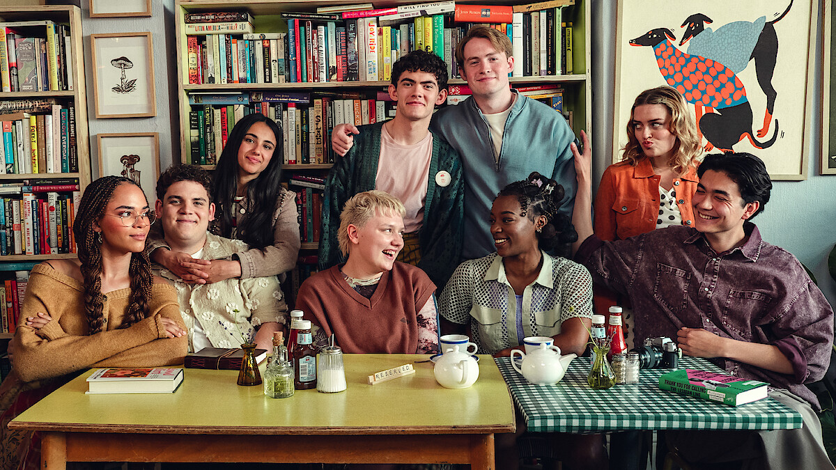 A diverse group of eight young people sitting and standing around a table in a cozy room with bookshelves and art, sharing a friendly moment, some smiling and talking, with tea and condiments on the table.