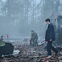 A man in a dark coat walks through debris with damaged buildings and rubble, while two people in camouflage uniforms investigate the scene. The area is foggy with leafless trees in the background, suggesting a disaster or crash site.
