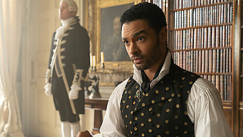 A man in Regency-era clothing sits in a richly decorated library with bookshelves and paintings, while a servant stands in the background near a window and candle holder.