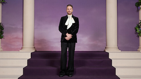 A man in a formal black suit stands on purple carpeted stairs between classical columns, in front of a purple backdrop with a soft sunset sky.