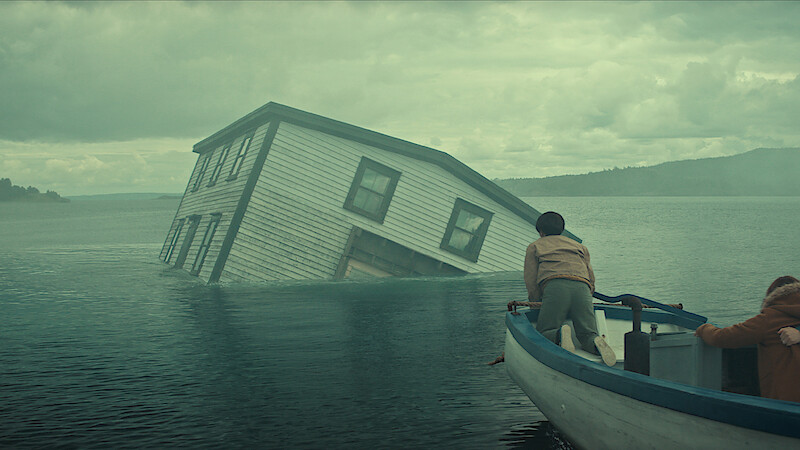 Two people in a small boat approach a partially submerged house tilted in the middle of a misty lake under a cloudy sky, with forested hills in the background.