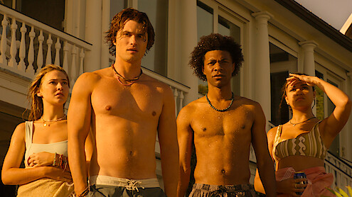 Four young adults in swimwear stand outside a large house, bathed in warm sunlight, looking intently ahead, suggesting a summer or beach setting and a moment of anticipation or concern.