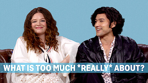 Megan Stalter and Will Sharpe sit against a blue seamless, smiling