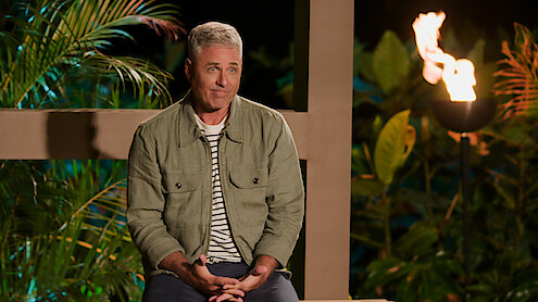 Mark L. Walberg sits outdoors at night, wearing a green jacket and striped shirt, surrounded by tropical plants and a lit torch in the background.
