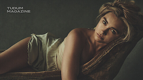 Woman with blonde hair in light clothing reclines on a vintage chair, looking directly at the camera in a dimly lit, moody setting. Tudum Magazine logo appears in the upper left corner.