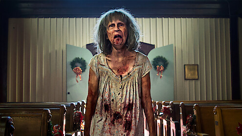 A disheveled, bloodied woman with gray hair stands in the aisle of a decorated church, with pews on either side and wreaths on the doors in the background, looking distressed or possibly zombified.