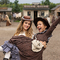 Two girls in brown vintage dresses and hats, smiling and posing playfully in a historic Western town setting with old buildings in the background and a dirt street.