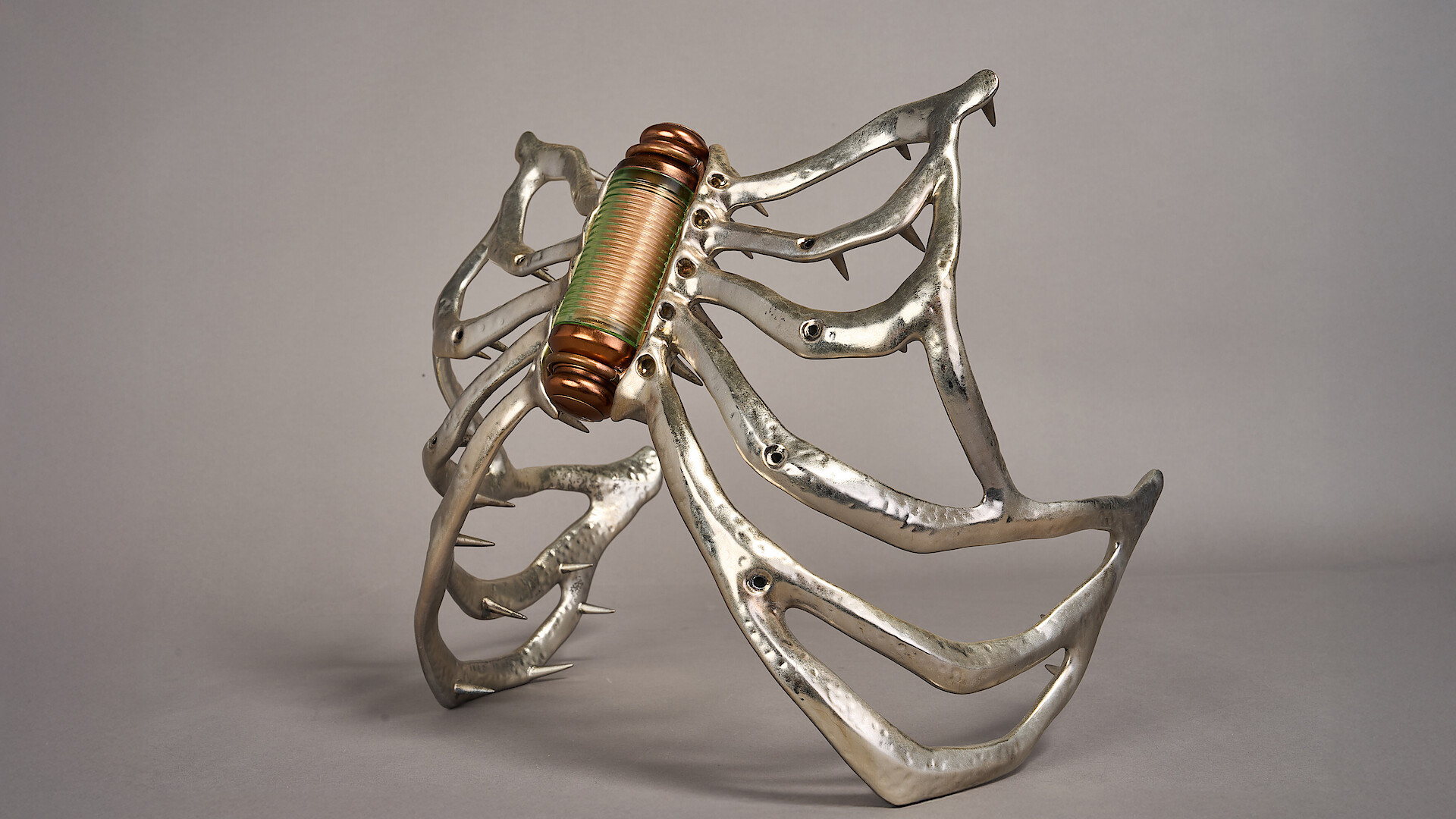 Futuristic spider-like sculpture with metallic legs and a copper coil body, set against a gray background. The mood is mechanical and surreal, evoking curiosity and intrigue.