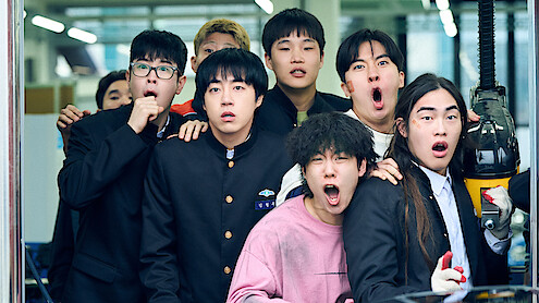 A group of surprised young men in school uniforms and casual clothes stand together indoors, looking in shock at something off-camera in a brightly lit modern setting.