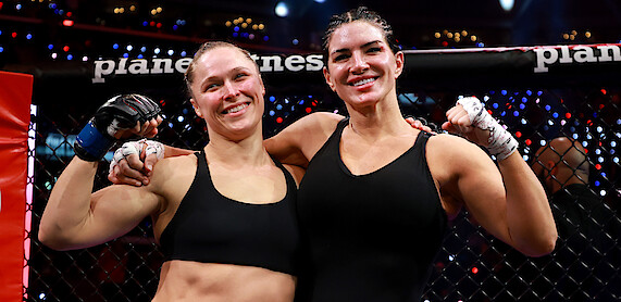 Two female MMA fighters in black sportswear stand arm-in-arm and smiling inside a caged arena after a match, with bright lights and cheering audience in the background.