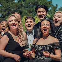 Group of young adults dressed in formal evening wear, laughing and smiling outdoors at a daytime event with trees in the background.
