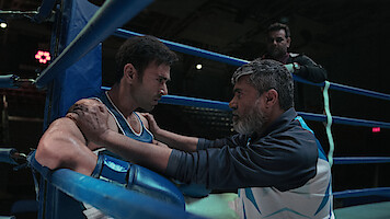 A boxer sits in a boxing ring corner, wearing blue and white gear, while his coach kneels in front of him giving advice; another person stands in the background, dimly lit arena.