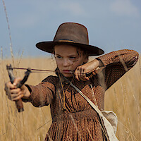 Young girl in a brown dress and wide-brimmed hat aims a slingshot in a field of tall dry grass under a blue sky, focusing intently.