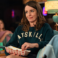 Woman sitting indoors on a couch wearing a "CATSKILLS" sweatshirt, casually eating from a red and white paper tray, with a vintage lamp and soft background lighting. Cozy, relaxed environment.