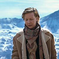 Person wearing a brown shearling coat and patterned scarf standing outdoors in a snowy mountain landscape on a sunny day.