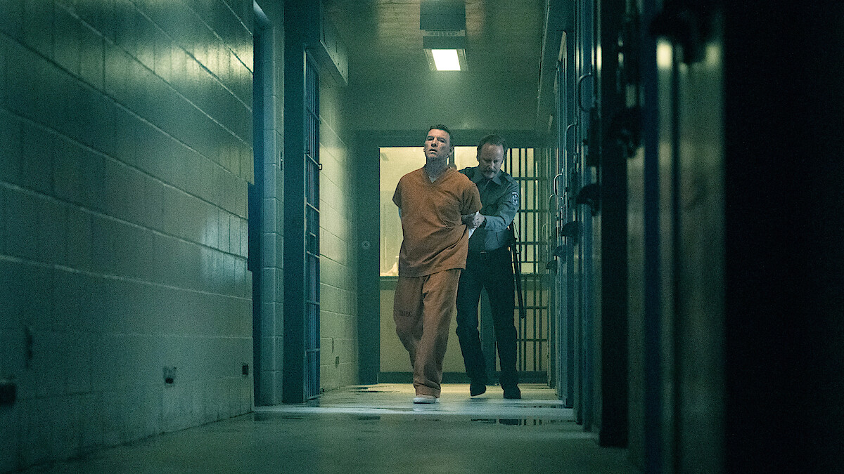 A man in an orange prison jumpsuit is being escorted by a guard down a dimly lit hallway in a jail or prison, with barred cells and concrete walls visible.