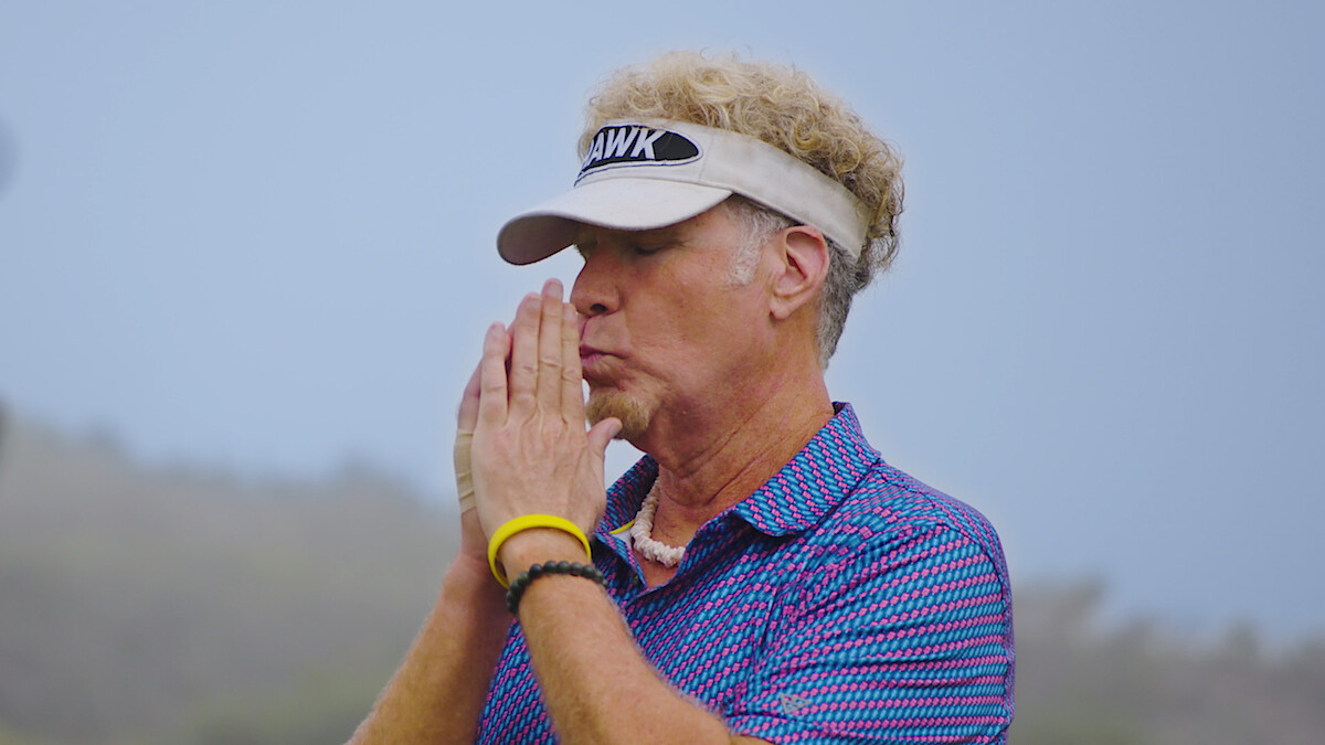 Man with curly blond hair in a visor stands outdoors, hands pressed together in front of face in a thoughtful or focused pose, wearing a blue patterned shirt, with a misty natural background.