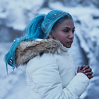 Charithra Chandran as Princess Vivi, with blue hair in a white winter coat stands outdoors in a snowy, icy landscape, looking thoughtful and cold with hands clasped, snowflakes in hair and on coat.