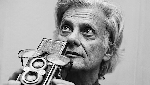 Older person with gray hair holding a vintage twin-lens reflex camera to their face, looking upward; indoor setting with a plain light background. Black and white photo.