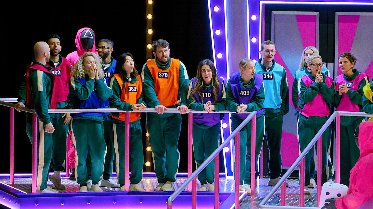 A group of contestants wearing numbered tracksuits stand on a brightly lit game show set, appearing tense and focused. Neon lights and a futuristic, colorful environment add excitement and intensity to the scene.