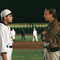 Two men face each other on a baseball field at night, one in a vintage baseball uniform and the other in casual clothes with a brown leather jacket, with a cornfield and players in the background.