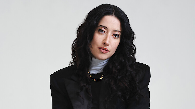 Taylor Ortega photographed in front of a white backdrop wearing a black jacket with strong shoulders, white turtle neck layered under a black shirt, and a gold chain with her dark curly hair cascading around her face.