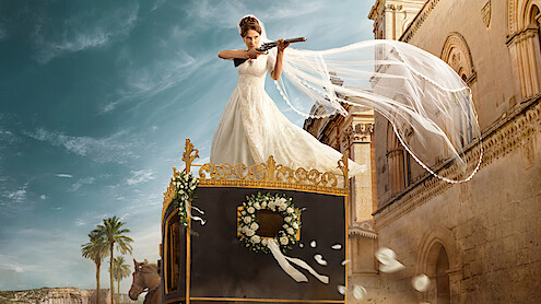 A bride in a white wedding dress stands on top of an ornate carriage, aiming a shotgun, with her veil flowing in the wind. The scene is set outdoors near classical buildings and palm trees under a bright sky.
