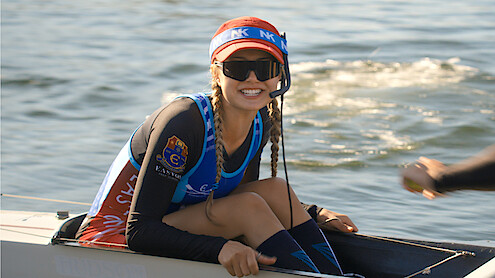 Smiling woman with braids wearing sunglasses, a visor, and athletic gear sits in a rowing boat on the water, holding equipment with one hand and a headset microphone, sunlight reflecting off the rippling water in the background.