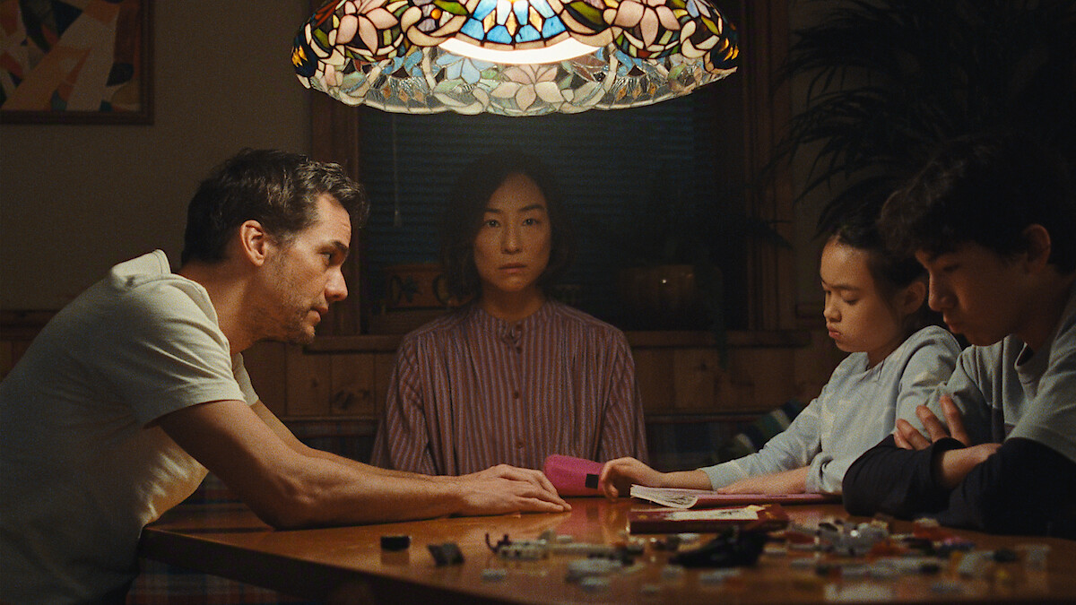 Four people sit around a wooden table under a stained glass lamp, focused on conversation and paperwork, in a cozy, warmly lit room with plants and wall art in the background.