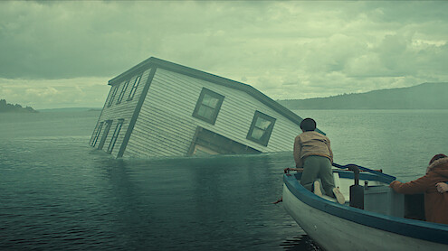 Two people in a small boat approach a partially submerged house tilted in the middle of a misty lake under a cloudy sky, with forested hills in the background.