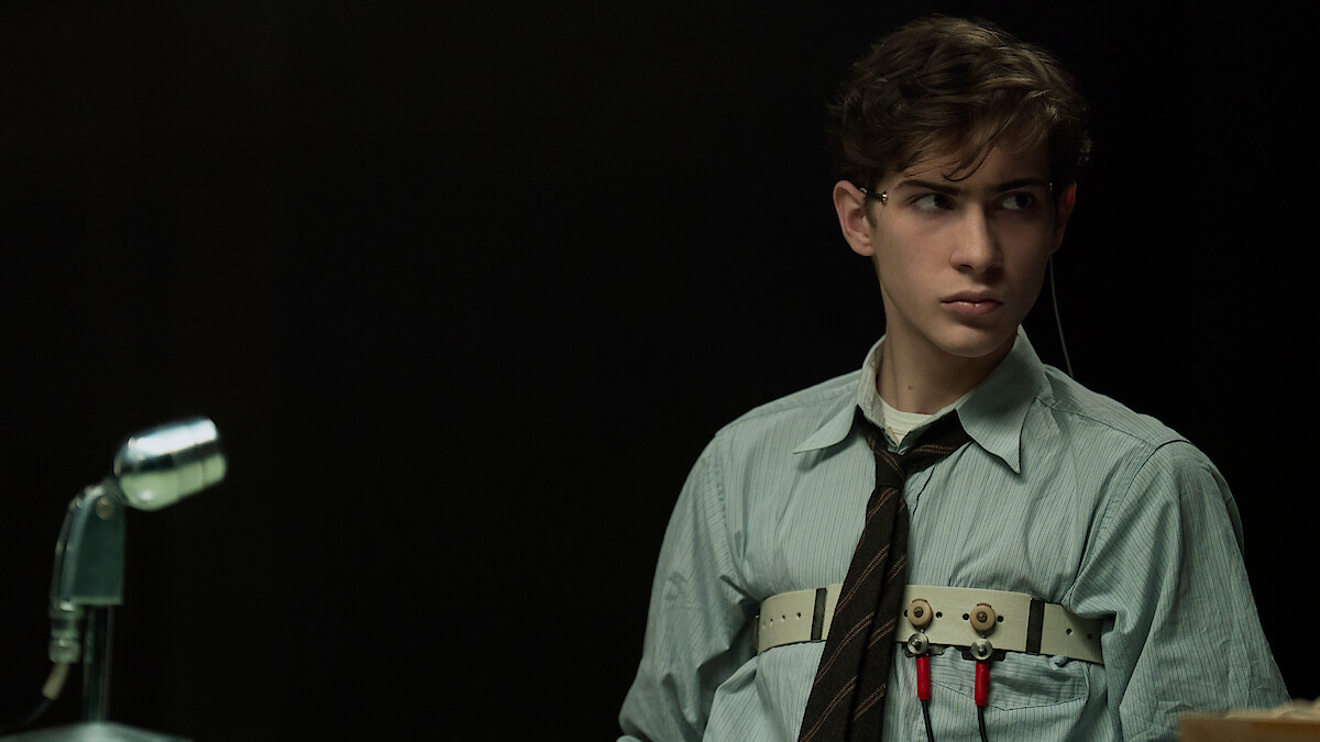 Young man in a collared shirt and tie sits in a dimly lit room, attached to a polygraph test device, with a microphone in
