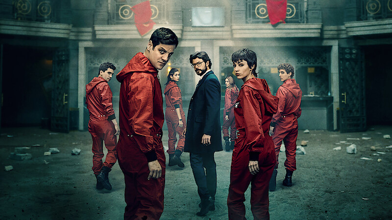 Group of people in red jumpsuits and a man in a suit stand in a dimly lit, grand building interior with balconies, flags, and scattered debris, suggesting a dramatic or tense atmosphere.