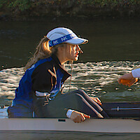 A female coxswain in a blue and black jacket and white cap sits in a rowing boat on the water, holding a microphone, with another rower’s hand visible, surrounded by natural outdoor scenery.